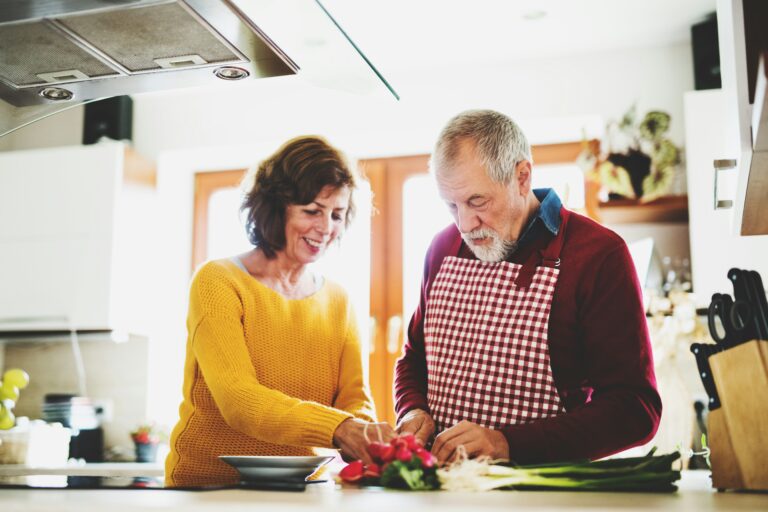 Casal preparando uma refeição na bancada da cozinha com um porta-temperos organizado ao lado.